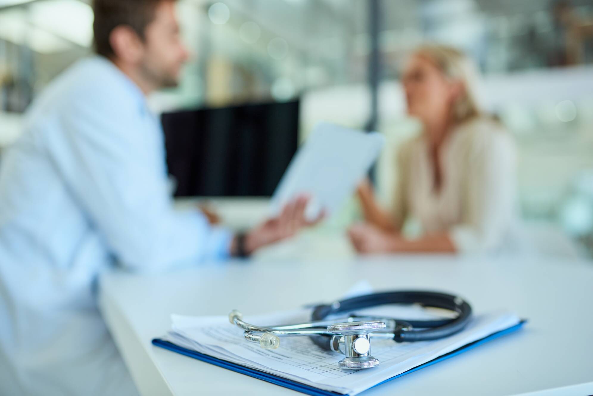 Foto de un estetoscopio y un portapapeles en un escritorio con un médico y un paciente en el fondo